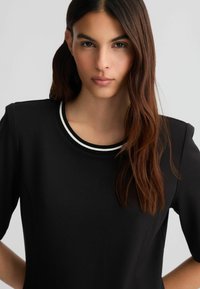 Abito nero con scollatura rotonda e doppia striscia bianca. Il tessuto è liscio, con maniche lunghe e una forma aderente.