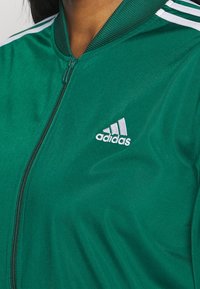Zielona kurtka Adidas na zamek z białymi paskami na ramionach i logo Adidas na lewej piersi, noszona przez osobę o ciemnych włosach.
