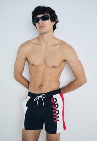 Crne kupaće gaće s bijelim i crvenim bočnim panelom na kojem je ispisano "HUGO". Gaće imaju pojas s vezicom i glatku teksturu.
