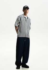 Short-sleeved, striped shirt in gray and white, paired with loose-fit, dark blue denim pants. Black shoes complete the outfit.
