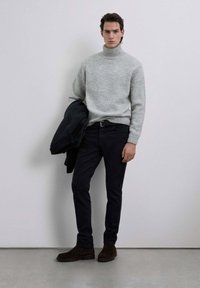 Suéter gris de cuello alto, jeans oscuros y botas de ante marrón. Modelo sosteniendo una chaqueta azul marino, posando frente a un fondo claro y sencillo.