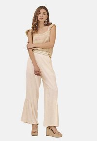 Pantalones acampanados de un beige claro combinados con una blusa sin mangas estampada que presenta rayas en tonos pastel. La modelo lleva sandalias de cuña y aretes largos.
