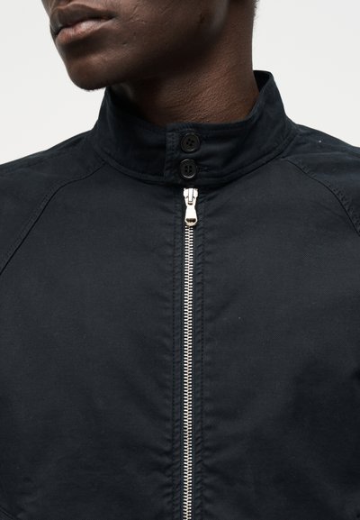 Κοντινό πλάνο ενός ατόμου που φοράει μαύρο μπουφάν με ψηλό γιακά, ασημένιο φερμουάρ και δύο μαύρα κουμπιά στον λαιμό.