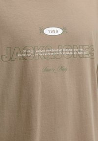 Bež majica s natpisom "JACK & JONES", ovalnim grbom "1990" i manjim tekstom koji se referira na urbanu streetwear estetiku i stil.