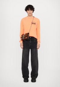 Neon orange langærmet skjorte med "Aries" logo, sorte bredde-ben jeans med sidesyninger, og en mørkebrun taske med kvægskindsdetaljer.