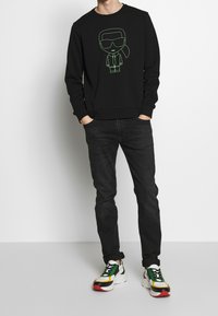 Felpa nera con un disegno a linea verde di un personaggio sul davanti, abbinata a jeans scuri e sneaker multicolori.