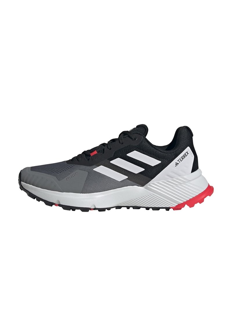 Zapatilla de trail running Adidas Terrex en negro y gris con franjas blancas y acentos rojos en la suela, vista de lado.