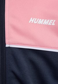Chaqueta deportiva de poliéster en rosa y azul marino con un logo blanco de "HUMMEL", costuras en contraste y cierre de cremallera.