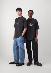 Černé trička s logem "Calvin Klein Jeans", kombinovaná s modrými cargo džíny a černými kalhotami, doplněná černými botami.