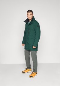 Grønn vanntett parkas med høy krage, glidelåslommer og svarte detaljer. Sammen med grå bukser og gule joggesko.