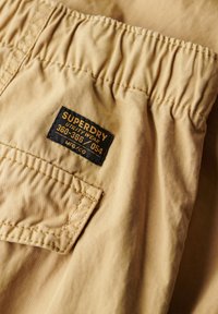 Shorts utilitaires beiges avec ceinture élastiquée, présentant une étiquette en tissu noir avec un texte jaune, et une poche arrière avec des détails cousus.