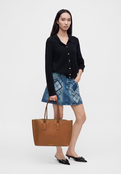 Femme portant un pull noir boutonné et une jupe en jean patchwork bleue, tenant un grand sac cabas en cuir marron avec des anses.