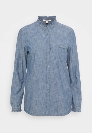 Blouse chambray bleu clair à manches longues avec col haut volanté, fermeture à boutons sur le devant, poche poitrine unique avec rabat et poignets volantés.