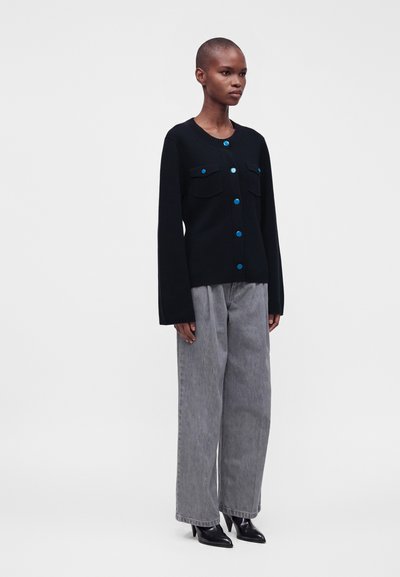 Femme aux cheveux courts portant un cardigan noir boutonné avec des boutons bleus, un jean gris à jambes larges et des bottes noires pointues, debout devant un fond uni.