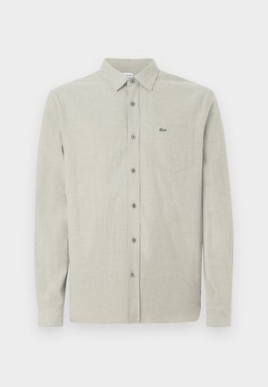 Camisa de manga larga color beige claro, con botón y cuello, bolsillo en el pecho que presenta un pequeño logo de cocodrilo verde.