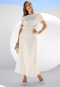 Cremefarbenes Maxikleid mit kurzen Ärmeln, elastischem Bund und seitlichem Schlitz. Gestylt mit goldenen Armreifen und einer beigen Clutch; kombiniert mit spitzen Absatzschuhen.