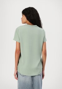 Blusa verde chiaro a maniche corte con scollatura rotonda, texture liscia e taglio dritto, caratterizzata da un orlo posteriore leggermente più lungo.