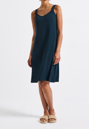 Kobieta ubrana w bezrękawnikową granatową sukienkę sięgającą kolan z marszczonymi ramiączkami i beżowymi plecionymi sandałami wsuwanymi, stojąca na białej podłodze.