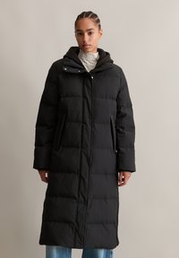 Vrouw in een lange zwarte pufferjas met capuchon, staande tegen een effen beige achtergrond, met spijkerbroek zichtbaar aan de onderkant.