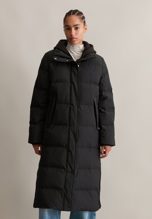 Vrouw in een lange zwarte pufferjas met capuchon, staande tegen een effen beige achtergrond, met spijkerbroek zichtbaar aan de onderkant.