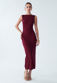 Burgundy sleeveless dress with a fitted silhouette, featuring a square neckline, smooth texture, and side slit. Worn with strappy gold sandals.
