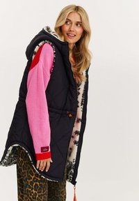 Frau trägt eine schwarze gesteppte Weste mit Kapuze und Leopardenmuster-Futter, einen rosa-roten Pullover und Leopardenmuster-Hose, schaut seitwärts und lächelt.