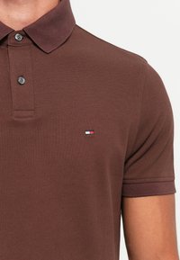 Nærbilde av overkropp iført brun poloskjorte med to knapper og lite rød, hvit og blått rektangulært logo på venstre bryst.