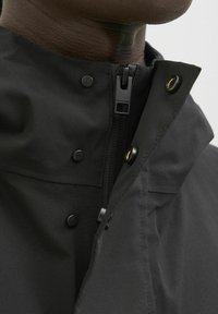 Jachetă neagră cu guler înalt, prevăzută cu închidere cu fermoar și butoane cu presiune. Material neted cu textură mată, branding subtil pe fermoar.