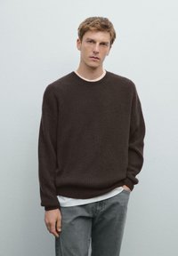 Donkerbruin geribbelde gebreide trui met een brede halslijn, gedragen over een wit T-shirt, gecombineerd met grijze jeans en de handen in de zakken.