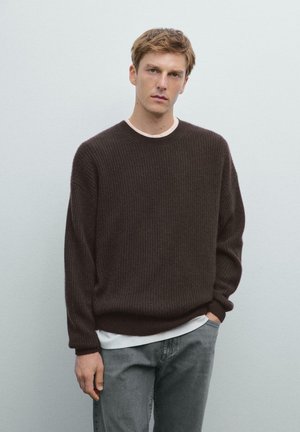 Donkerbruin geribbelde gebreide trui met een brede halslijn, gedragen over een wit T-shirt, gecombineerd met grijze jeans en de handen in de zakken.