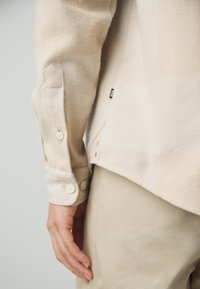 Poignet de chemise beige à manches longues avec boutons et petite étiquette "BOSS", portée avec un pantalon beige assorti sur le bras et la main d'une personne.