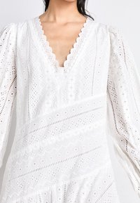 Abito bianco in pizzo ad occhio di pernice con scollatura a V profonda, bordi ondulati, maniche lunghe a palloncino e motivi sovrapposti con dettagli a taglio.