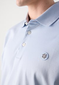 Polo azul claro feito de tecido suave, apresentando um colarinho, três botões e um emblema redondo com um logotipo prateado no peito.