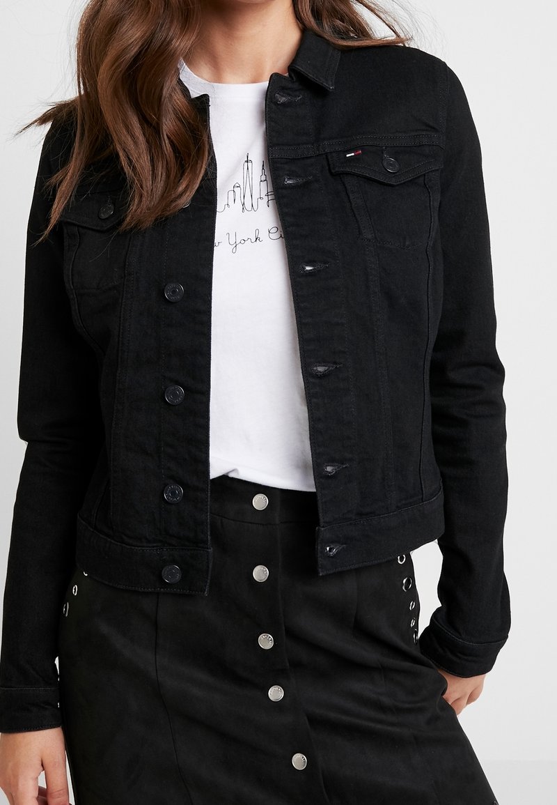 Woman wearing a black denim jacket over a white t-shirt with a New York City skyline sketch and a black buttoned skirt with silver details.