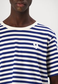 Camiseta de rayas con franjas horizontales azules y blancas, cuello redondo, y un pequeño logo de flor blanca en la parte izquierda del pecho. Material de algodón.