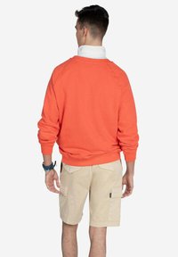 Sudadera naranja brillante de manga larga, con puños acanalados y cuello redondo. Combinada con pantalones cortos de carga beige que tienen bolsillos laterales.