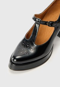 Chaussure en cuir noir avec un bout pointu, un design découpé et une sangle avec boucle. Présente un motif perforé décoratif sur le devant.