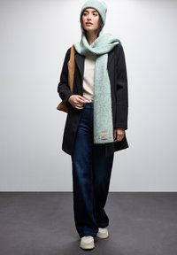 Sort frakke, creme sweater, mørkeblå brede bukser, lysegrøn tørklæde, matchende hue, lysebrun taske, og beige sneakers, på en grå baggrund.