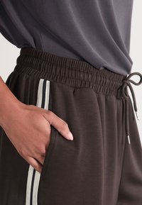 Braune Jogginghose mit elastischem Bund, Kordelzug und Seitentaschen. Weiße und marineblaue vertikale Streifen akzentuieren die Seiten.