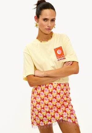 Femme portant des boucles d'oreilles en forme d'étoile, un t-shirt jaune "Le Soleil" et une jupe à motifs rouge, rose et jaune avec un ourlet à franges, bras croisés.