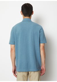 Short-sleeve polo shirt in light blue textured fabric, featuring a flat collar and a straight cut, with subtle variations in color throughout.