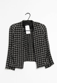 Czarna i biała kurtka typu blazer o kroju cropped w deseń houndstooth, bez kołnierza, z zamykanymi kieszeniami i zwisającą etykietą. Teksturowana tkanina.