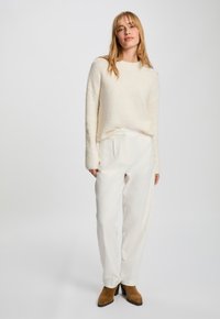 Kermavärinen tekstuurimainen neule, jossa on pitkät hihat, yhdistetty korkeavyötäröisiin valkoisiin housuihin ja ruskeisiin korkeakorkoisiin nilkkabootseihin. Yksinkertainen, minimalistinen tyyli.