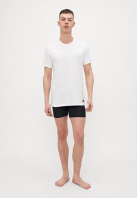 Λευκό βαμβακερό t-shirt με στρογγυλή λαιμόκοψη, σε συνδυασμό με μαύρες βερμούδες. Το μοντέλο στέκεται ξυπόλητο, αναδεικνύοντας την εφαρμογή και τον απλό σχεδιασμό του ρούχου.