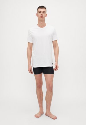 T-shirt bianca in cotone con scollo rotondo, abbinata a pantaloni corti neri. Il modello sta in piedi a piedi nudi, evidenziando la vestibilità e il design semplice del capo.
