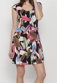 Femme portant une robe noire à manches courtes avec des motifs de feuilles colorées en rouge, vert, bleu et beige, des boutons sur le devant, debout devant un fond uni.