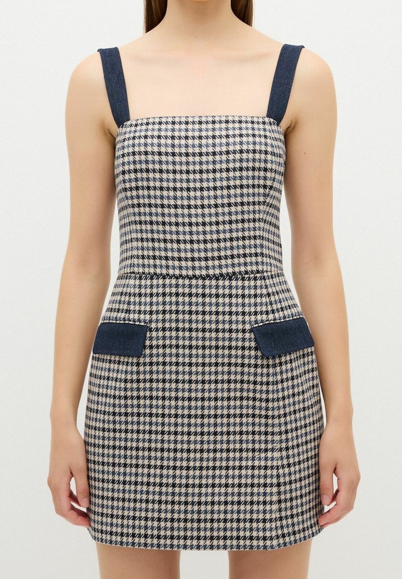 Femme portant une robe sans manches à motif pied-de-poule longueur genou avec des bretelles bleu foncé et des rabats de poche assortis sur un fond uni.
