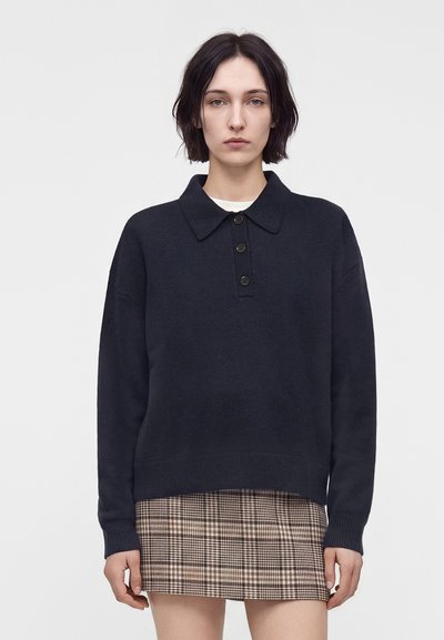 Jeune femme aux cheveux courts et foncés portant un pull noir à col et une mini-jupe marron à carreaux, debout devant un fond clair uni.