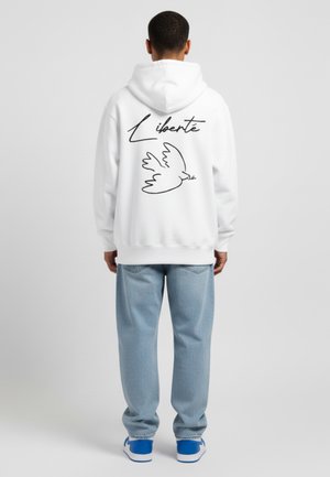 Un hoodie blanc avec le mot "Liberté" en lettre cursive noire et un dessin d'une colombe au dos. Associé à un jean bleu clair et des baskets bleues.
