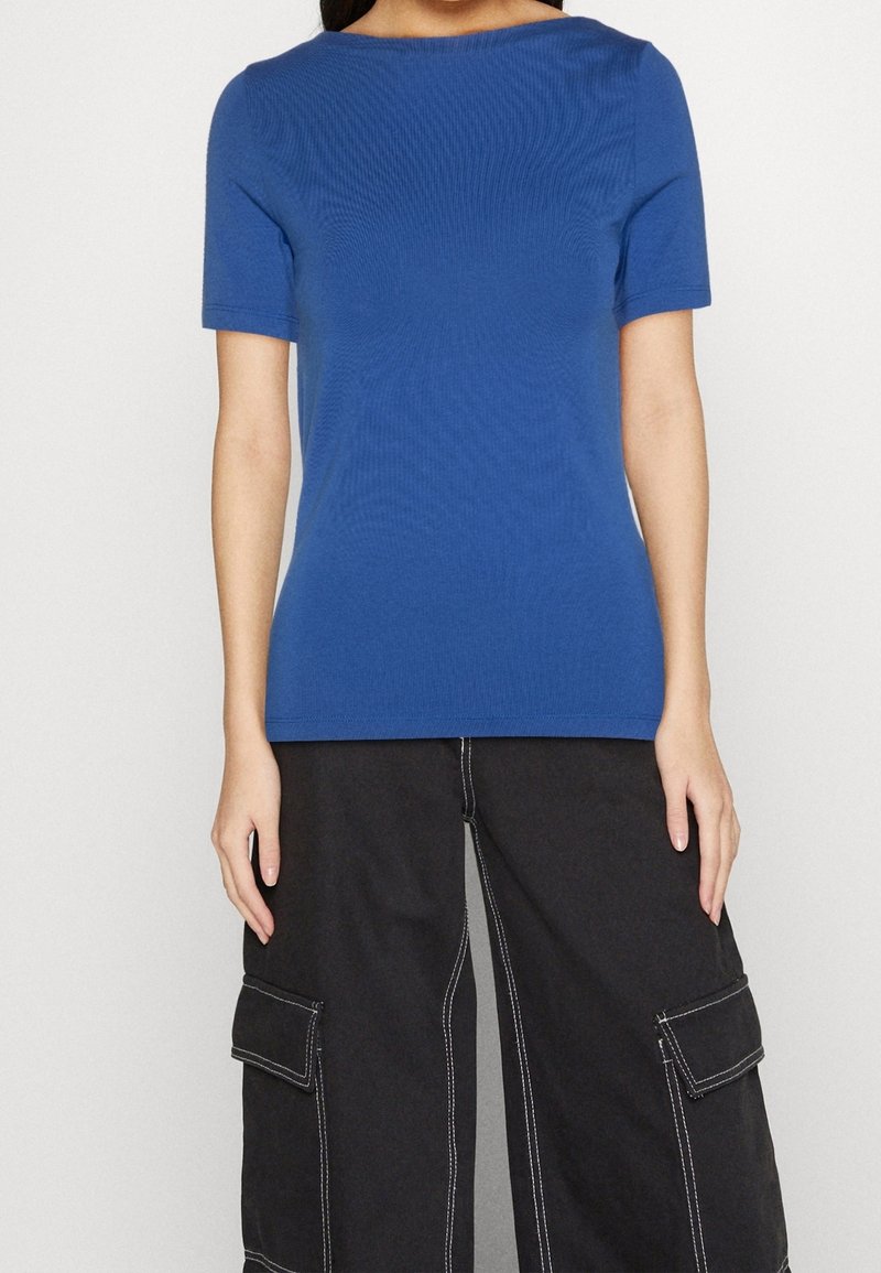 Personne portant un haut bleu à manches courtes ajusté et un pantalon cargo noir large avec des coutures blanches sur un fond blanc uni.